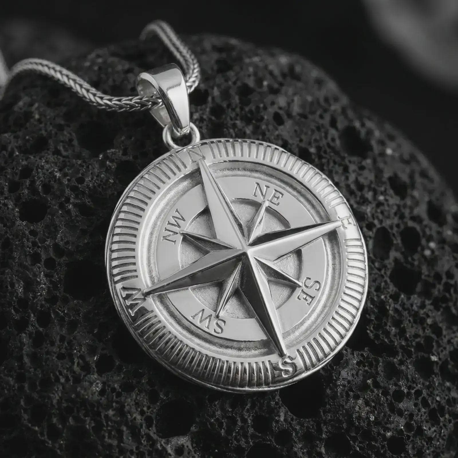 Polished silver compass pendant necklace on textured volcanic rock, centered macro jewelry photo with detailed navigation markings, sharp star relief, and bright silver highlights.