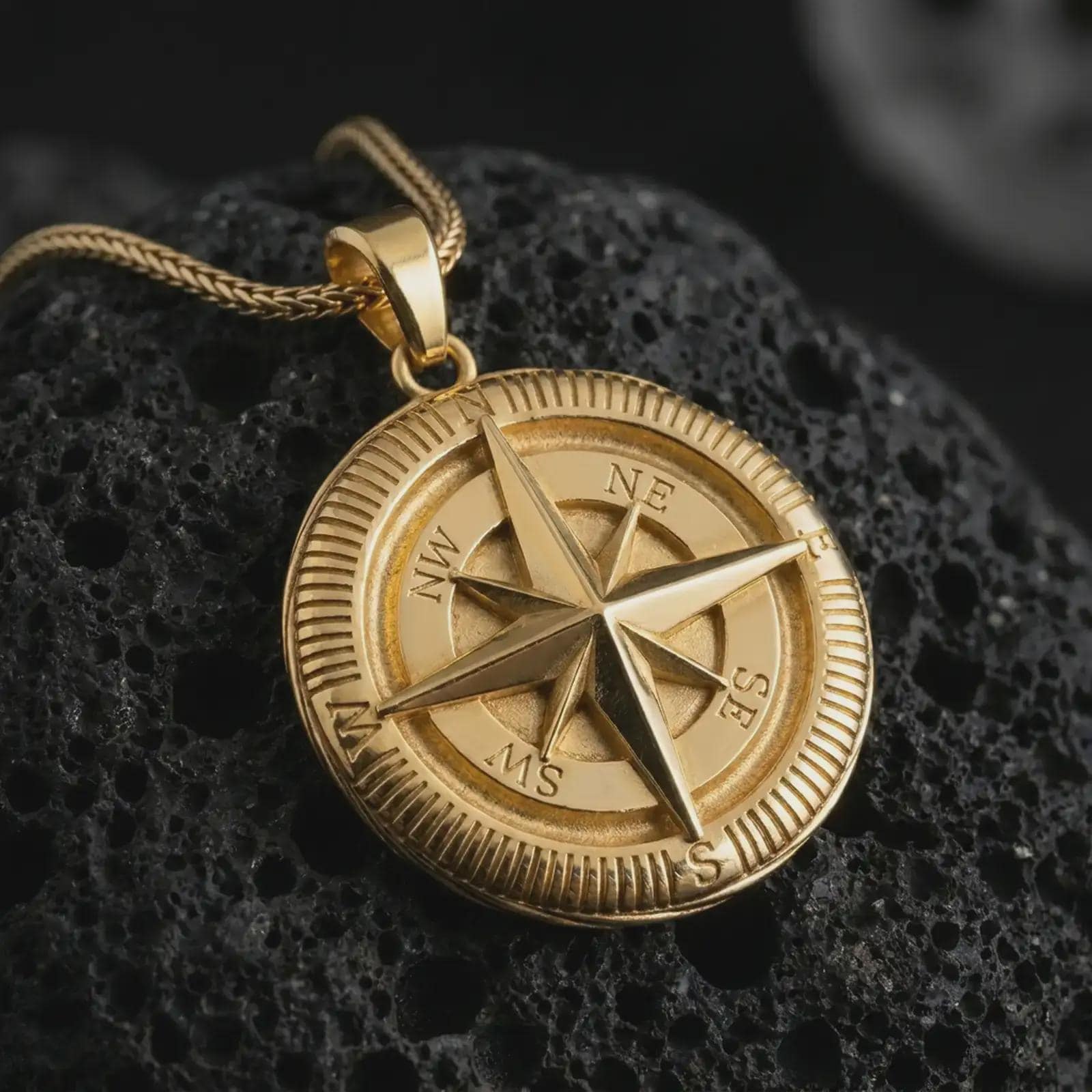Gold plated compass pendant necklace on textured volcanic rock, premium close-up studio composition emphasizing the nautical amulet design, cardinal directions, and explorer symbolism.