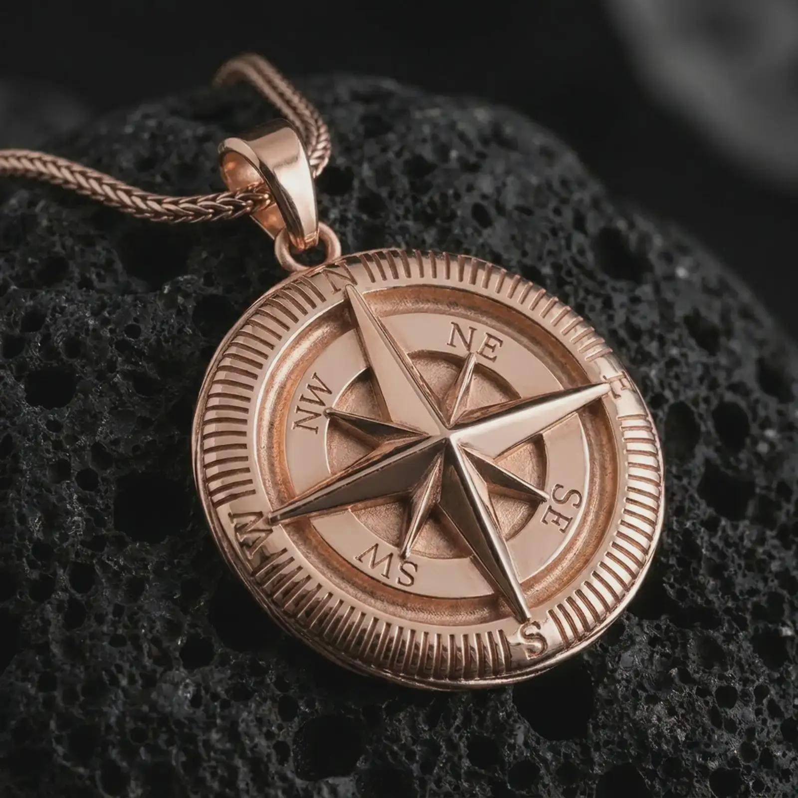 Rose gold plated compass pendant necklace on textured volcanic rock, detailed macro product shot with soft warm reflections and symbolic journey-inspired styling.