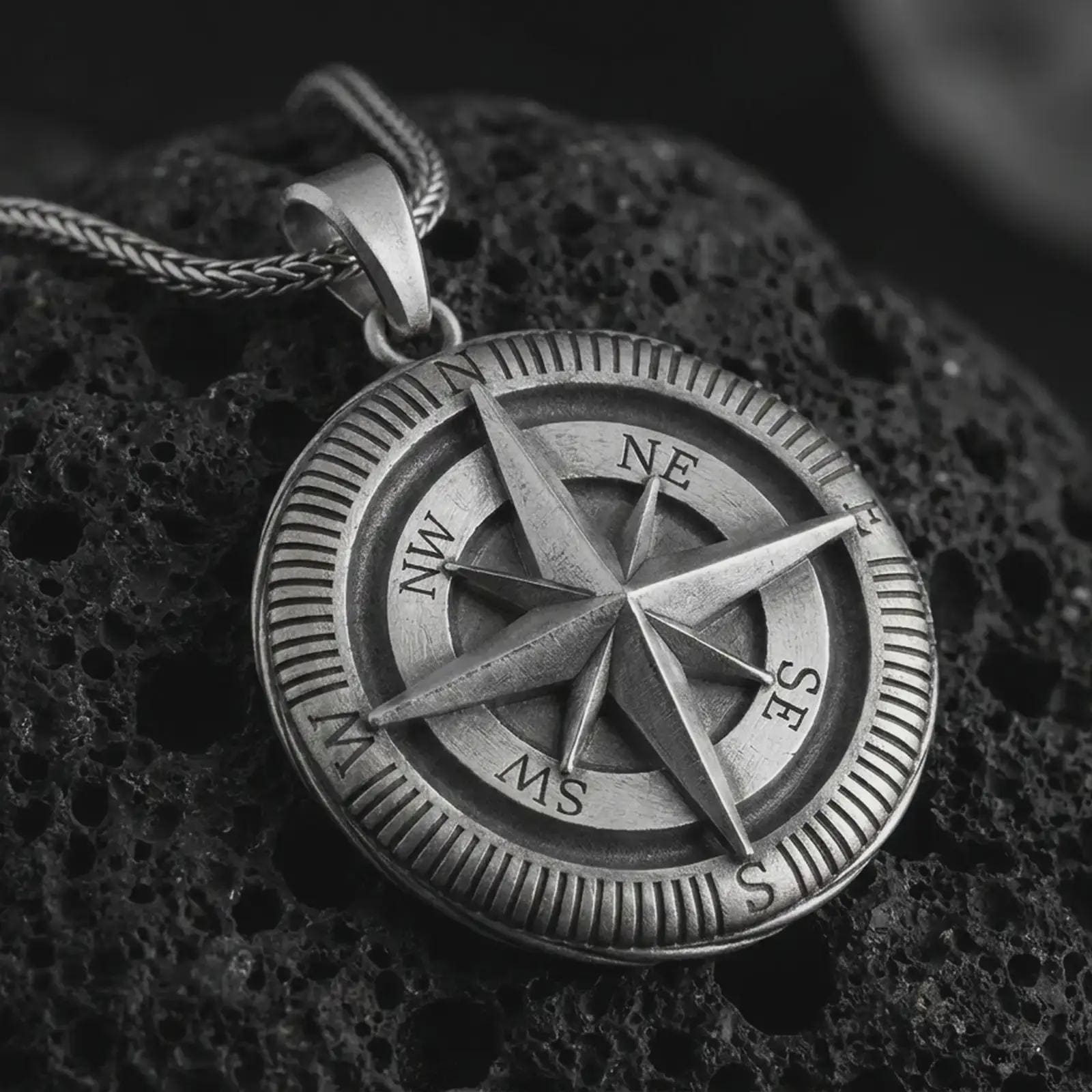 Oxidized compass pendant necklace on textured volcanic rock, close-up studio shot highlighting the raised compass rose, directional letters, ridged outer border, and antique silver contrast.
