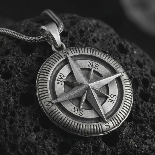Oxidized compass pendant necklace on textured volcanic rock, close-up studio shot highlighting the raised compass rose, directional letters, ridged outer border, and antique silver contrast.