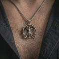 Load image into Gallery viewer, Oxidized Vitruvian Man pendant necklace worn on the chest, close-up lifestyle jewelry photo showing the Leonardo da Vinci art pendant on a spiga chain with dark clothing.
