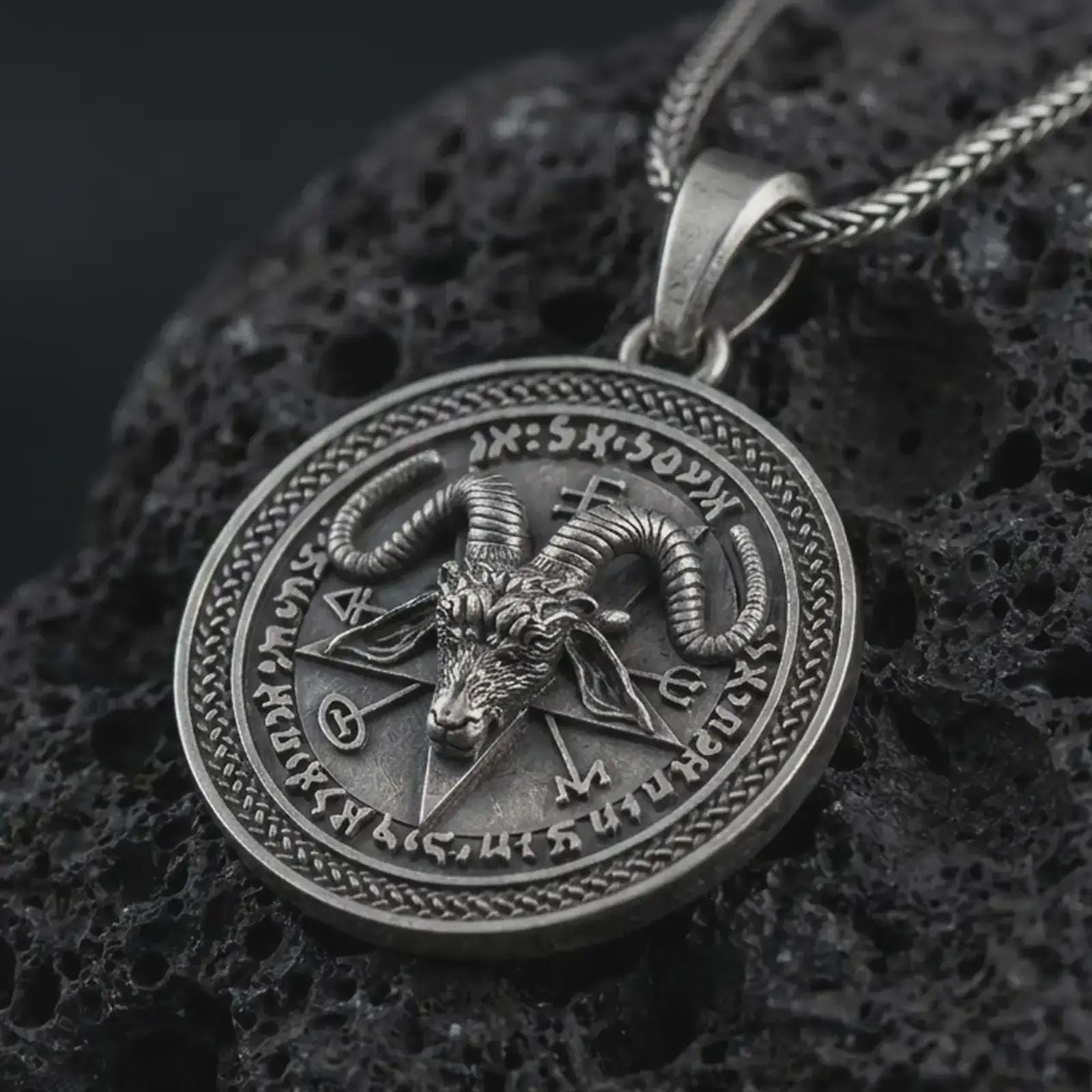 Oxidized Baphomet pendant necklace on textured volcanic rock, close-up studio shot highlighting the goat head relief, pentagram background, occult sigils, and antique silver contrast.