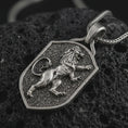 Load image into Gallery viewer, Oxidized heraldic lion shield pendant necklace on textured volcanic rock, close-up studio shot highlighting the rampant lion relief, shield frame, textured background, and antique silver contrast.
