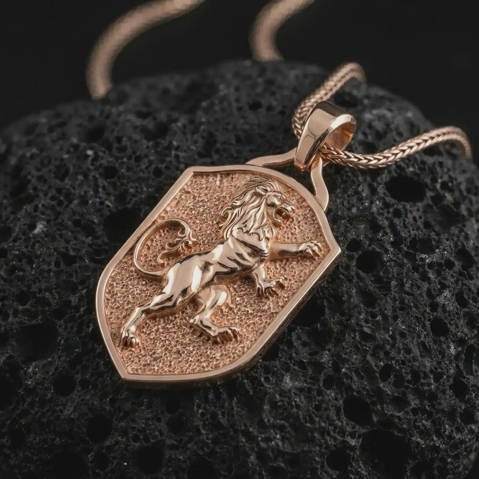 Rose gold plated heraldic lion shield pendant necklace on textured volcanic rock, detailed macro product shot with soft warm reflections and symbolic lion crest styling.