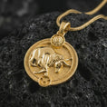 Load image into Gallery viewer, Gold plated Taurus zodiac pendant necklace on textured volcanic rock, premium close-up studio composition emphasizing the bull figure, sun motif, and astrology medallion design.
