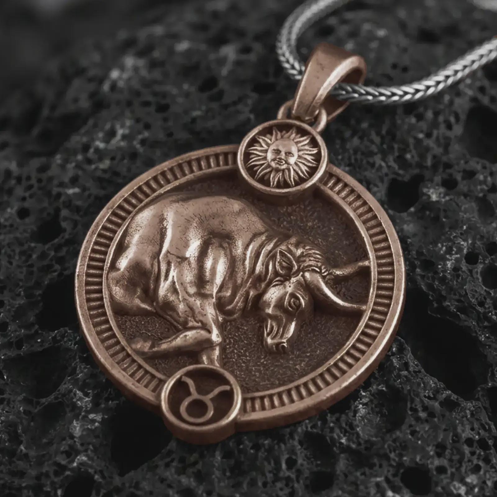 Bronze Taurus zodiac pendant necklace on textured volcanic rock, close-up studio shot highlighting the bull relief, sun detail, Taurus symbol, and antique silver contrast.
