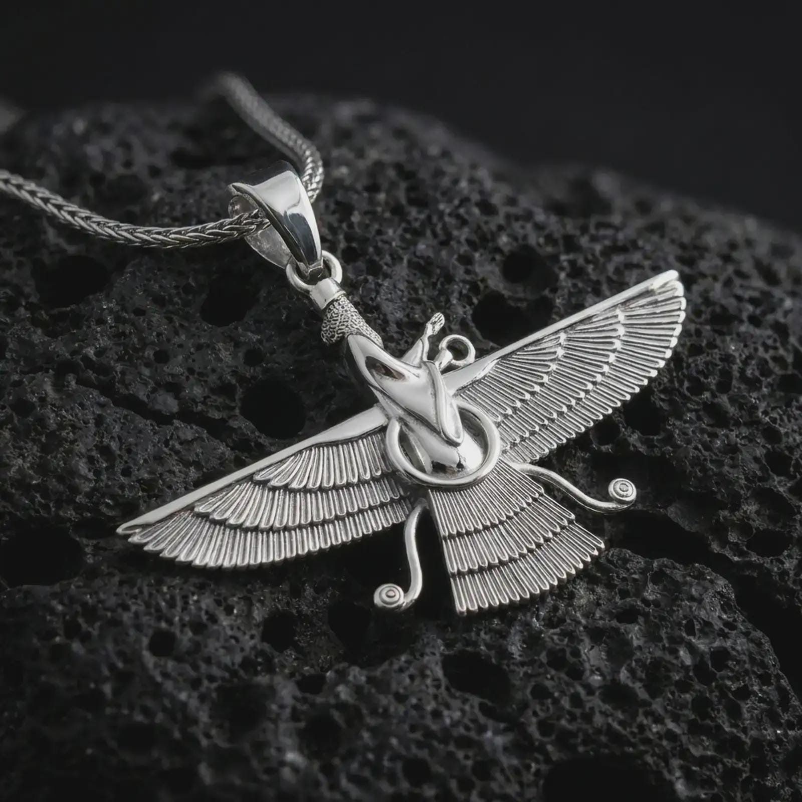 Polished silver Faravahar pendant necklace on textured volcanic rock, centered macro jewelry photo with detailed Ahura Mazda-inspired relief and bright silver highlights.
