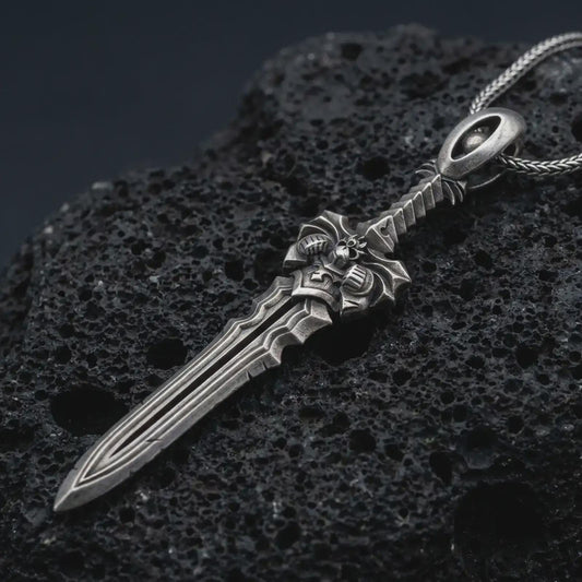An oxidized fantasy sword pendant rests on dark volcanic rock in a macro studio close-up, showing a detailed warrior blade with gothic sculptural elements.