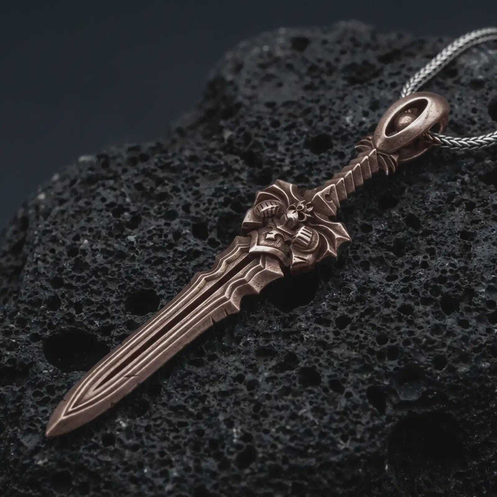 A Bronze fantasy sword pendant rests on dark volcanic rock in a macro studio close-up, showing a detailed warrior blade with gothic sculptural elements.