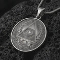 Load image into Gallery viewer, Oxidized all-seeing eye pendant necklace on textured volcanic rock, close-up studio shot highlighting the triangle eye symbol, laurel wreath, radiant background, and antique silver contrast.
