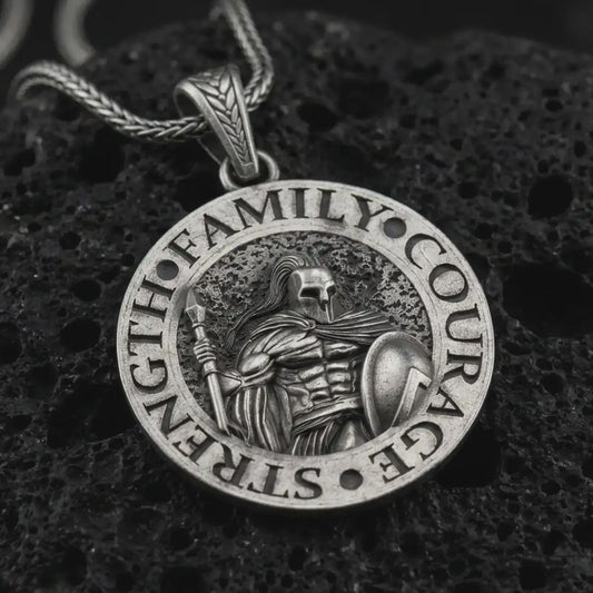Oxidized Spartan warrior pendant necklace on textured volcanic rock, close-up studio shot highlighting the helmeted soldier, spear, shield, strength lettering, and antique silver contrast.