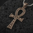 Load image into Gallery viewer, Bronze ankh cross pendant necklace on textured volcanic rock, close-up studio shot highlighting the ancient Egyptian key of life symbol, hieroglyphic details, and antique silver contrast.

