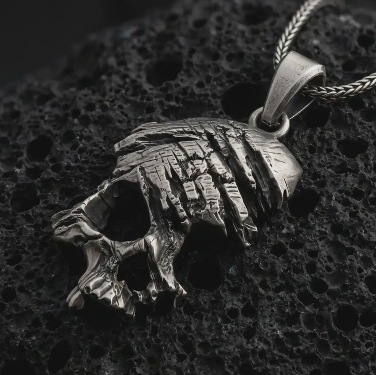 Oxidized cracked skull pendant necklace on textured volcanic rock, close-up studio shot highlighting the fractured skull form, deep cut details, gothic texture, and antique silver contrast.