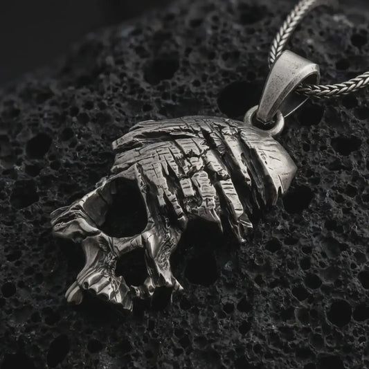 Oxidized cracked skull pendant necklace on textured volcanic rock, close-up studio shot highlighting the fractured skull form, deep cut details, gothic texture, and antique silver contrast.
