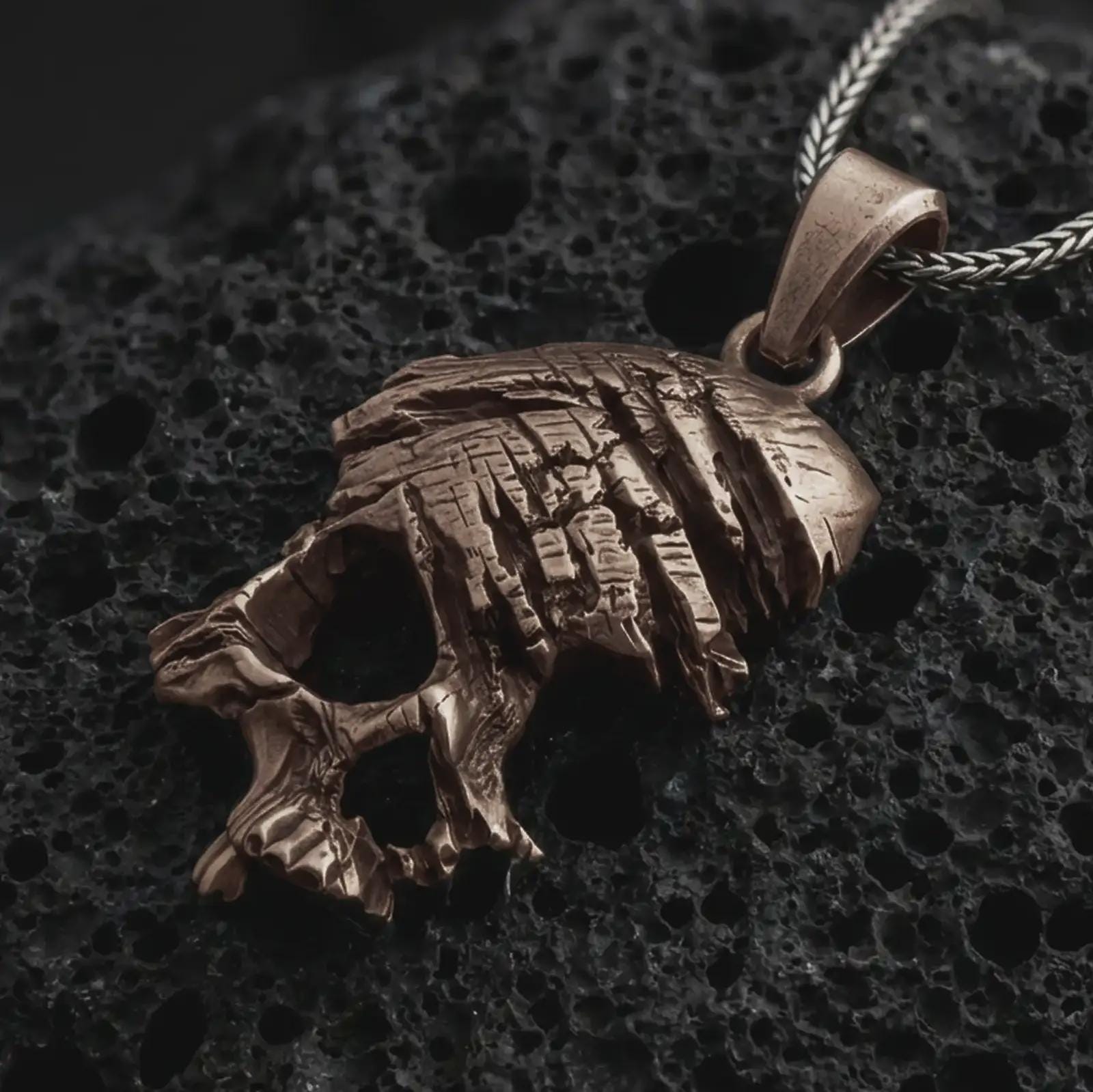 Bronze cracked skull pendant necklace on textured volcanic rock, close-up studio shot highlighting the fractured skull form, deep cut details, gothic texture, and antique silver contrast.