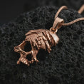 Load image into Gallery viewer, Rose gold plated cracked skull pendant necklace on textured volcanic rock, detailed macro product shot with soft warm reflections and bold gothic skull styling.
