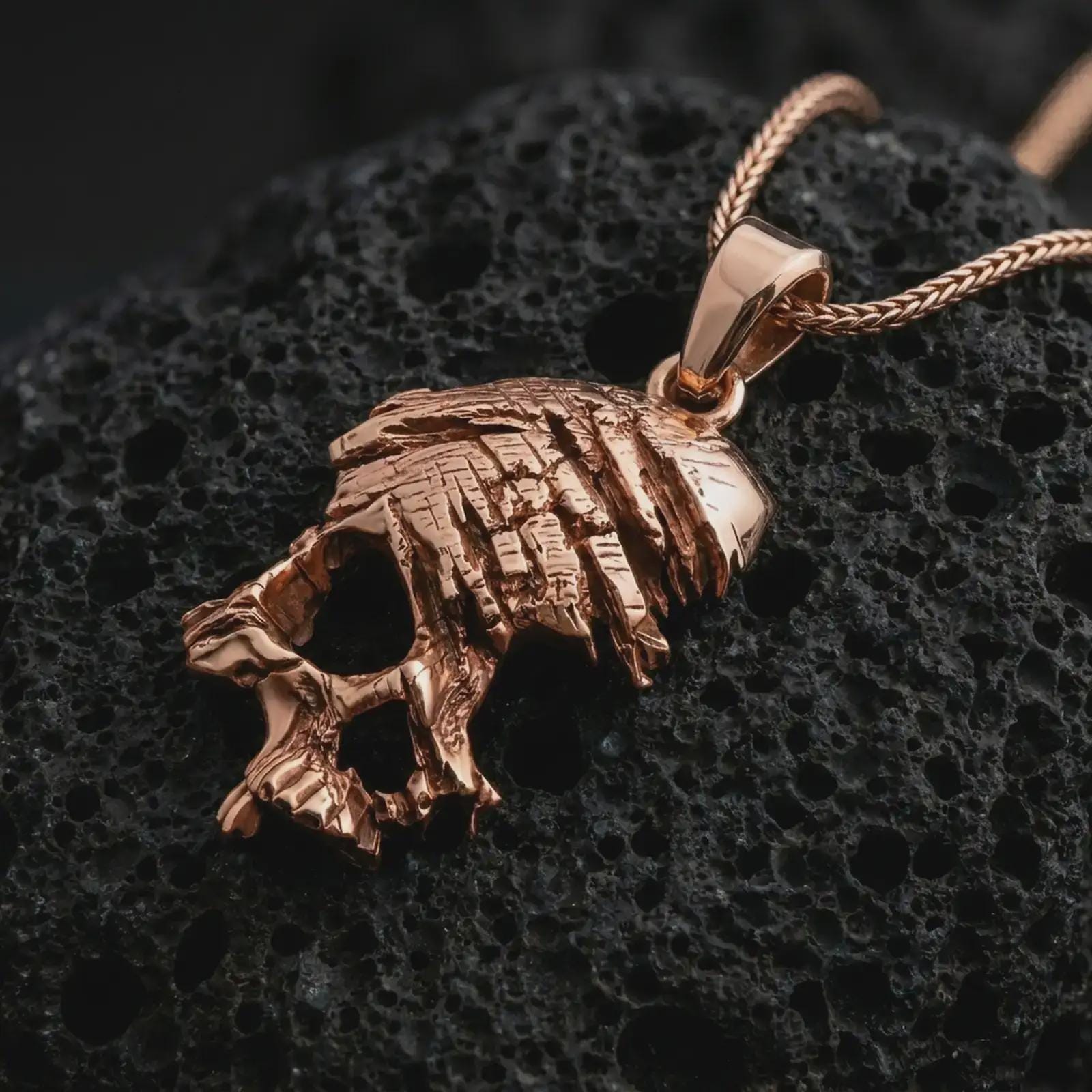 Rose gold plated cracked skull pendant necklace on textured volcanic rock, detailed macro product shot with soft warm reflections and bold gothic skull styling.