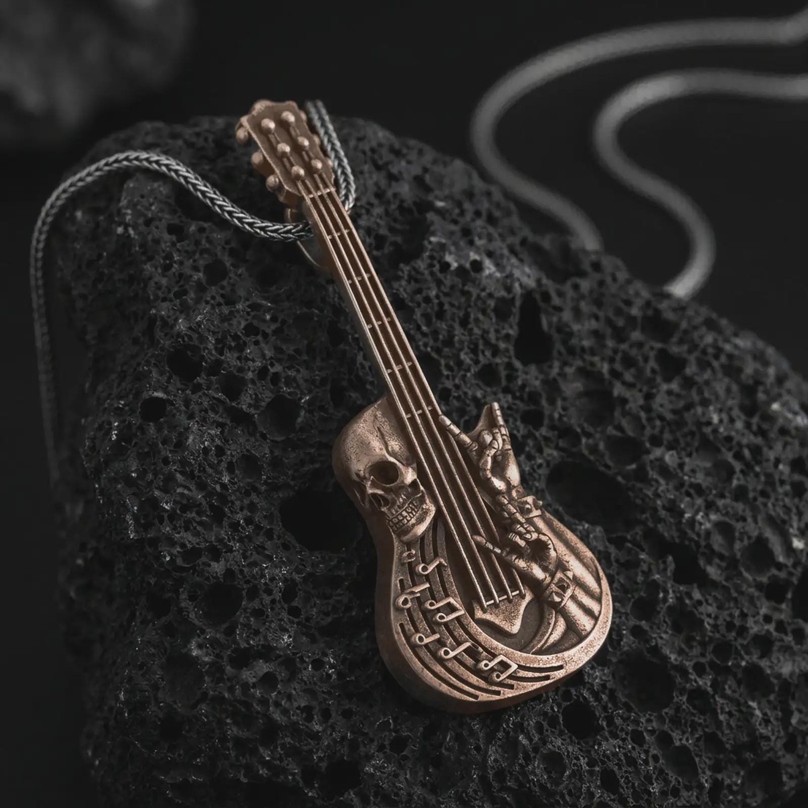 Bronze skull guitar pendant necklace on textured volcanic rock, close-up studio shot highlighting the guitar body, skull detail, rock hand gesture, music notes, and antique silver contrast.