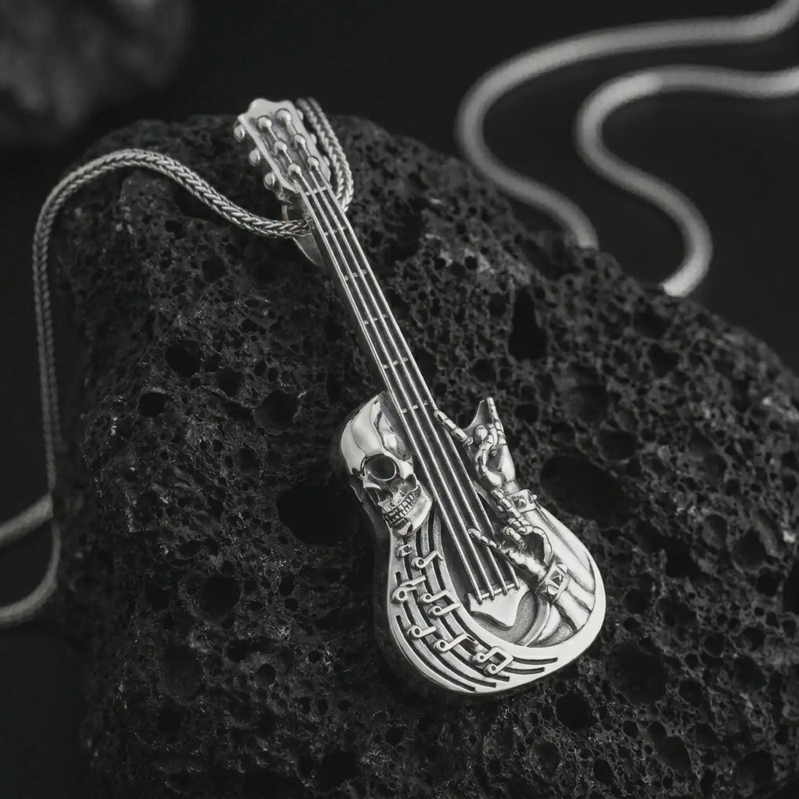 Polished silver skull guitar pendant necklace on textured volcanic rock, centered macro jewelry photo with detailed gothic musician relief and bright silver highlights.