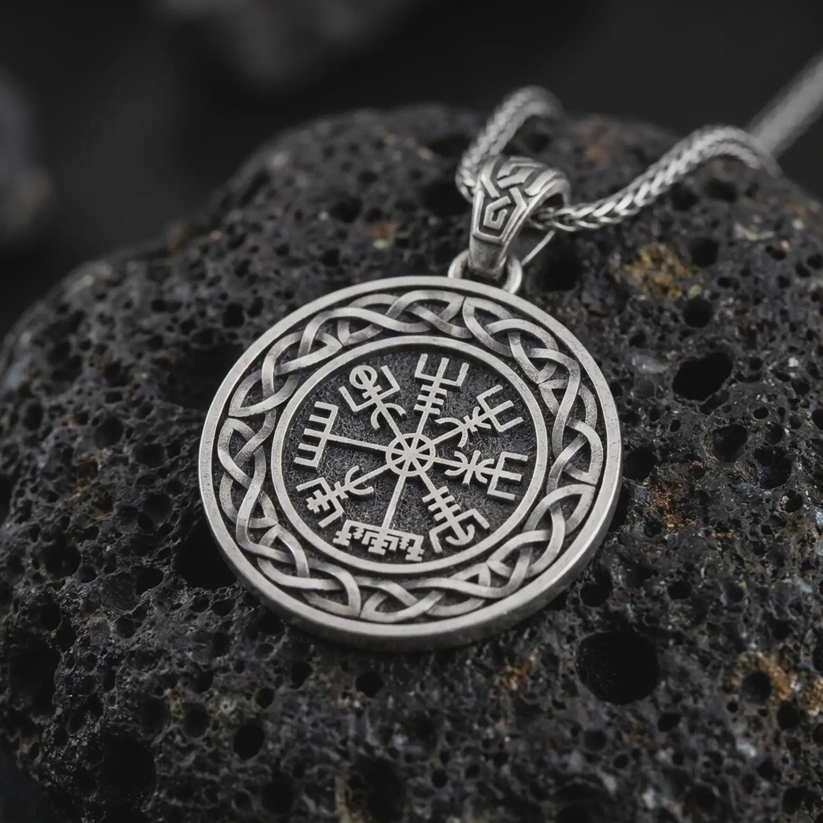 Oxidized Vegvisir Viking compass pendant necklace on textured volcanic rock, close-up studio shot highlighting the Norse protection symbol, Celtic knot border, and antique silver contrast.