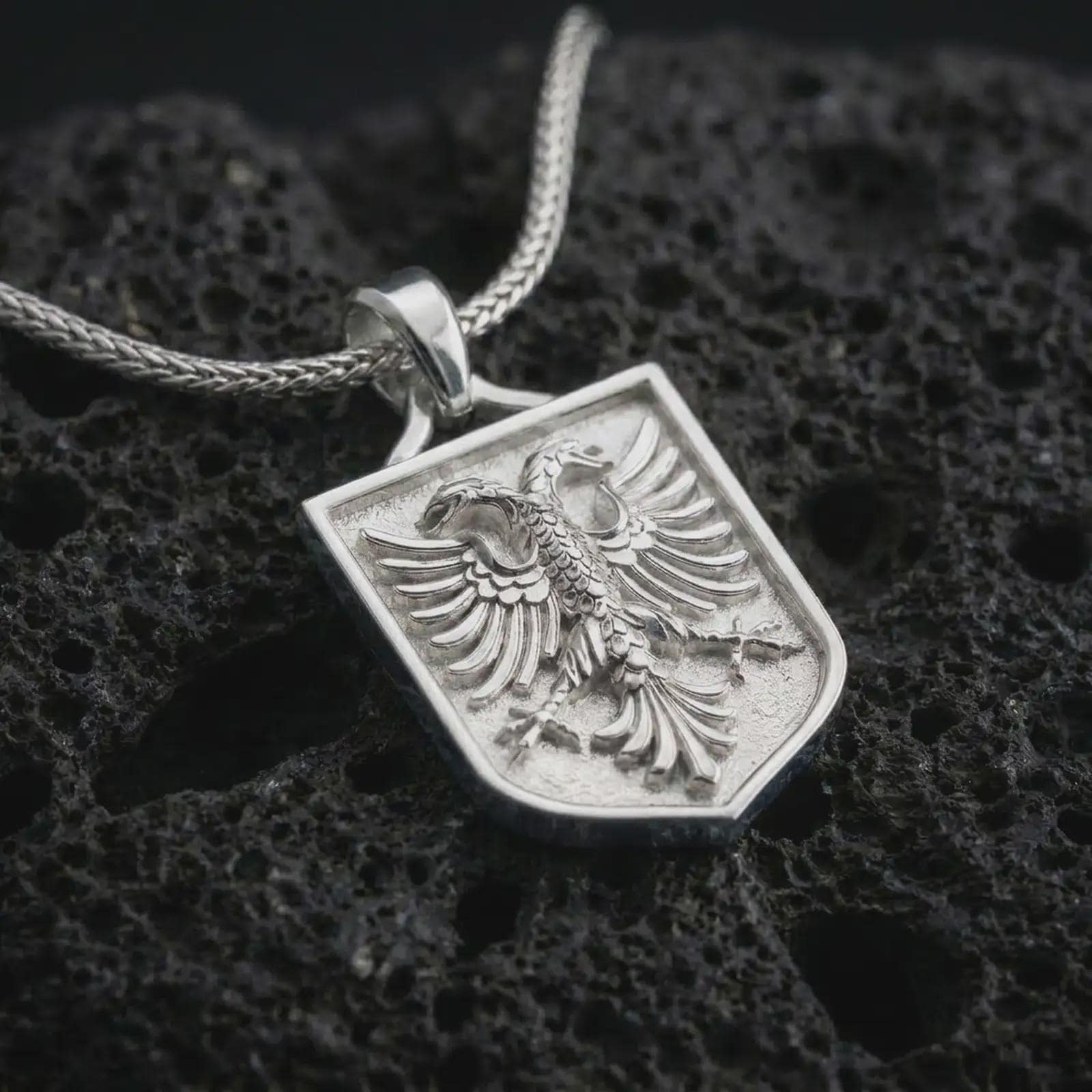 Polished silver double-headed eagle shield pendant necklace on textured volcanic rock, centered macro jewelry photo with detailed feather relief and bright silver highlights.