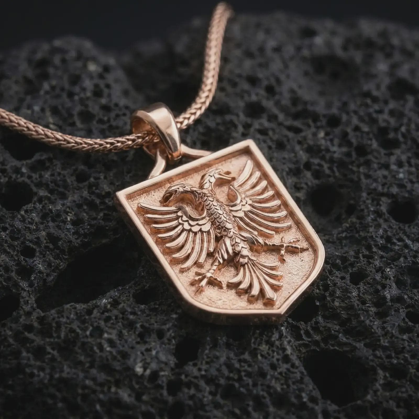 Rose gold plated double-headed eagle shield pendant necklace on textured volcanic rock, detailed macro product shot with soft warm reflections and symbolic Balkan eagle styling.