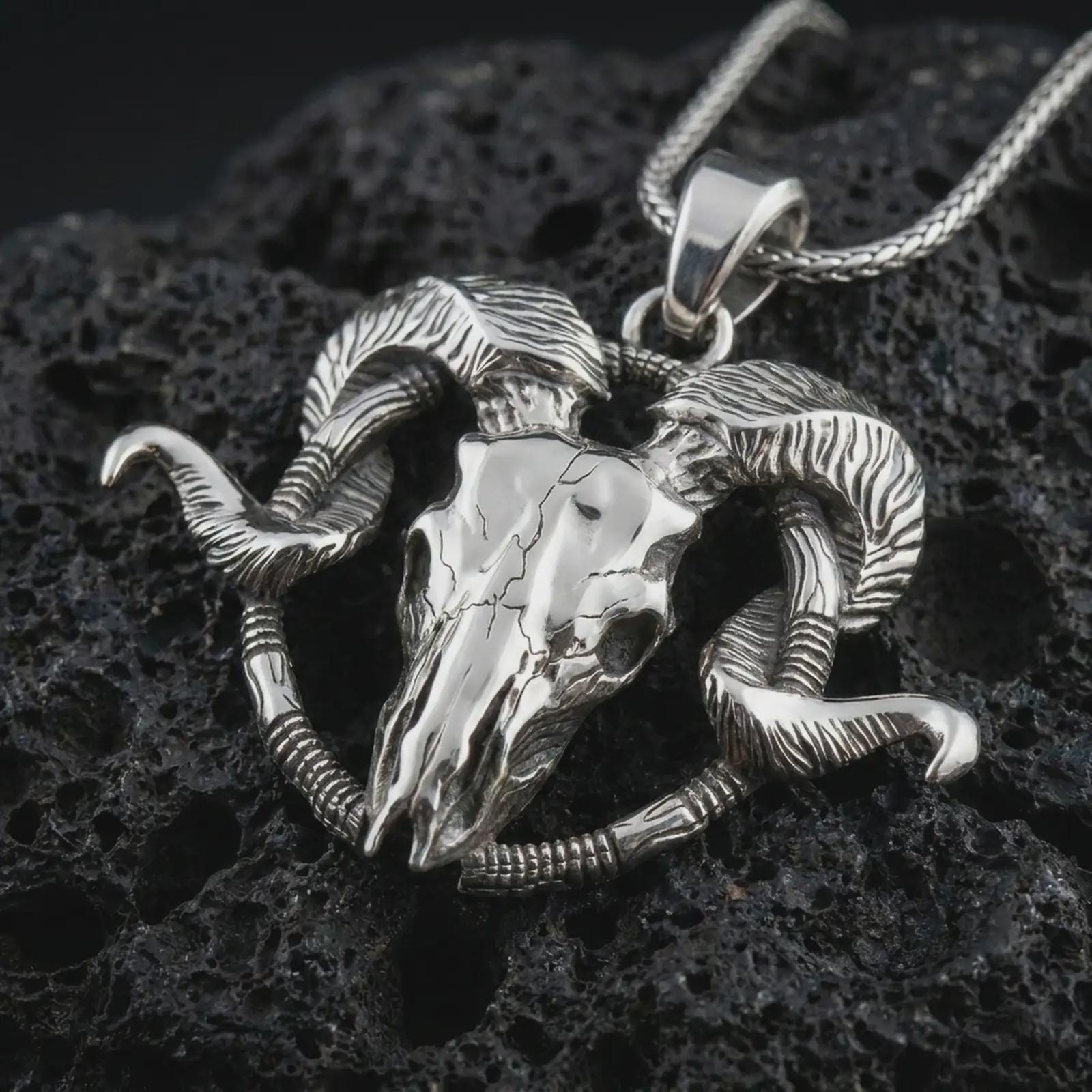 Polished silver ram skull pendant necklace on textured volcanic rock, centered macro jewelry photo with detailed horn engraving, skull texture, and bright silver highlights.