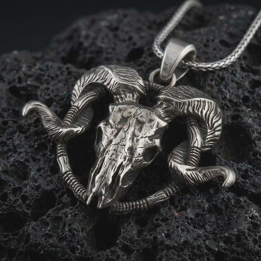 Oxidized ram skull pendant necklace on textured volcanic rock, close-up studio shot highlighting the cracked skull, curled horns, skeletal details, and antique silver contrast.