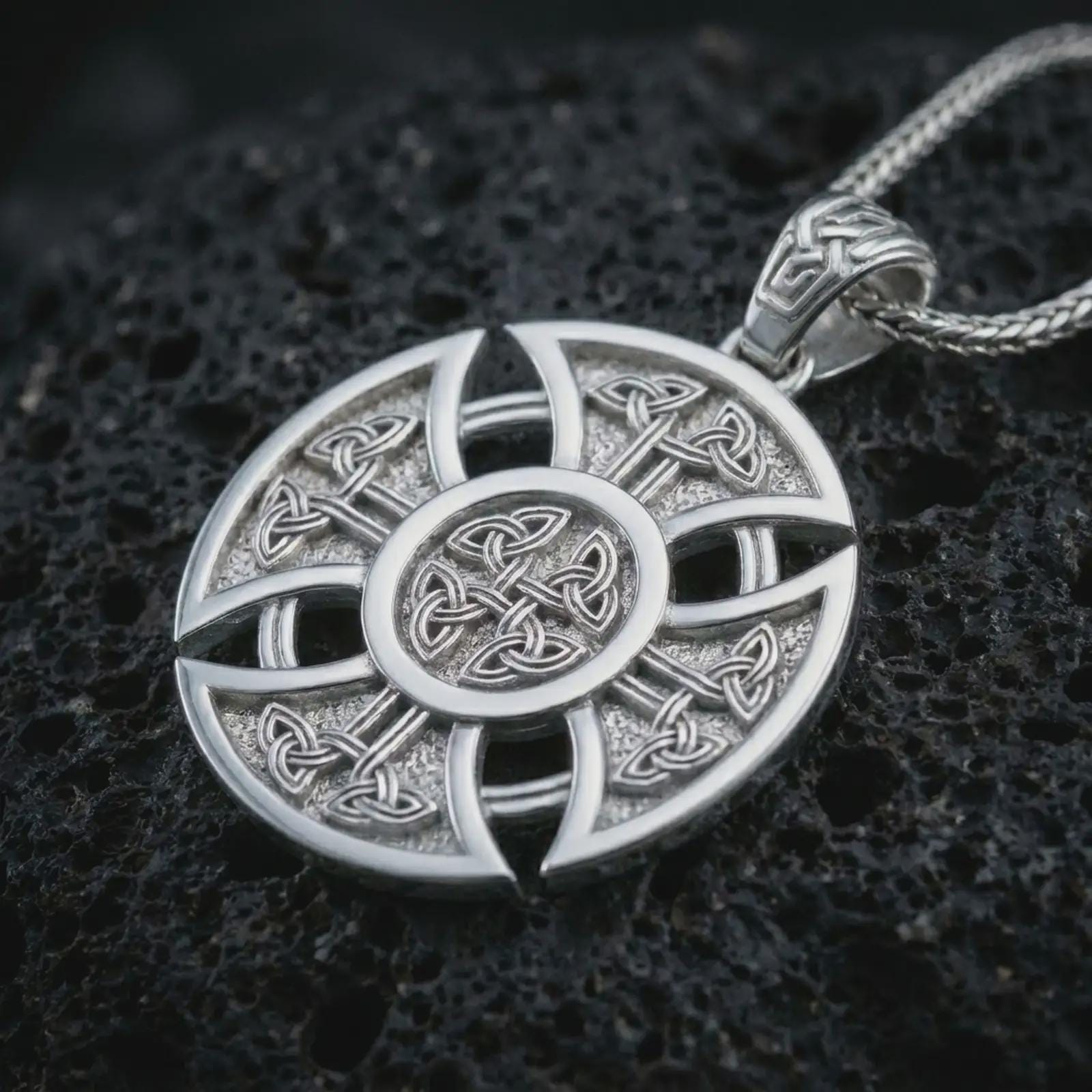Polished silver Celtic cross pendant necklace on textured volcanic rock, centered macro jewelry photo with detailed knotwork engraving and bright silver highlights.
