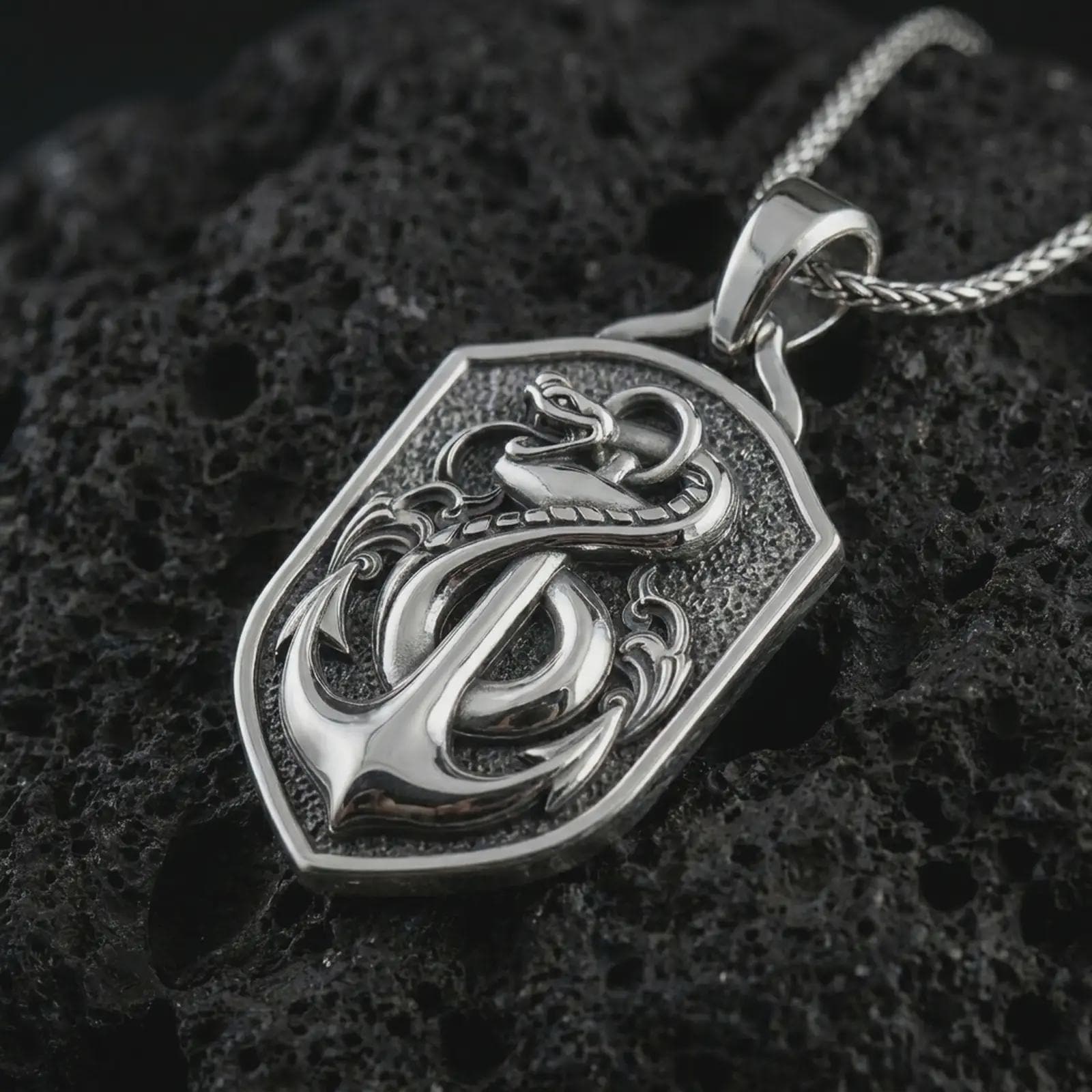 Polished silver serpent and anchor pendant necklace on textured volcanic rock, centered macro jewelry photo with detailed maritime relief and bright silver highlights.