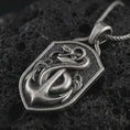 Load image into Gallery viewer, Oxidized serpent and anchor pendant necklace on textured volcanic rock, close-up studio shot highlighting the coiled snake, nautical anchor, shield frame, and antique silver contrast.
