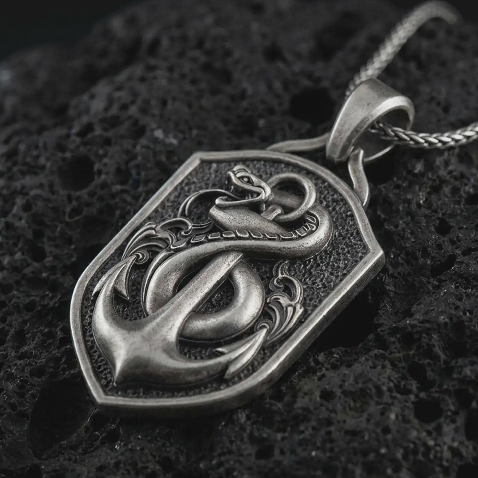 Oxidized serpent and anchor pendant necklace on textured volcanic rock, close-up studio shot highlighting the coiled snake, nautical anchor, shield frame, and antique silver contrast.