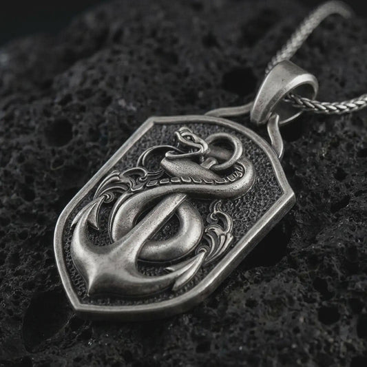 Oxidized serpent and anchor pendant necklace on textured volcanic rock, close-up studio shot highlighting the coiled snake, nautical anchor, shield frame, and antique silver contrast.