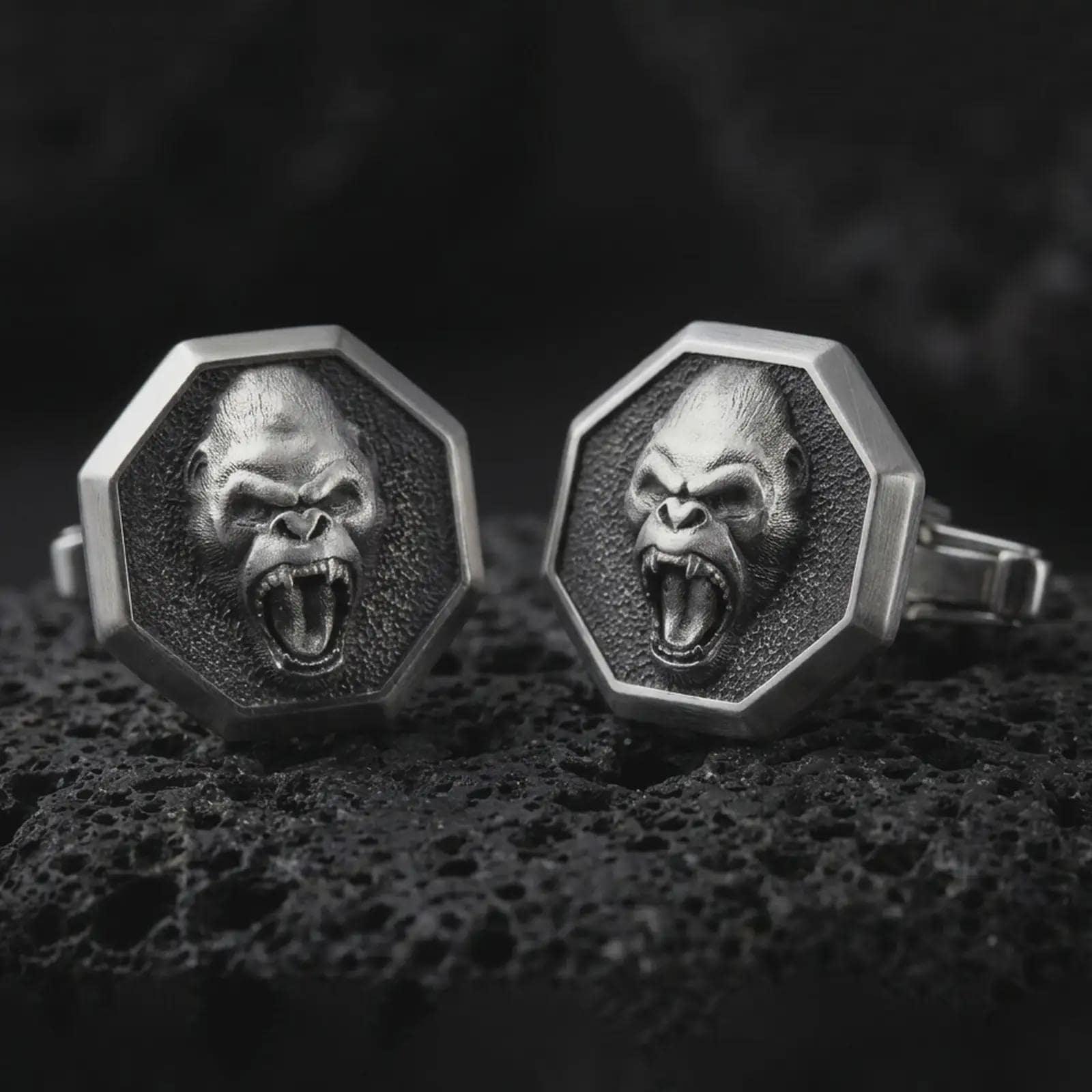 Oxidized gorilla cufflinks on textured volcanic rock, close-up studio shot highlighting the roaring ape face, octagonal frame, deep relief, and antique silver contrast.