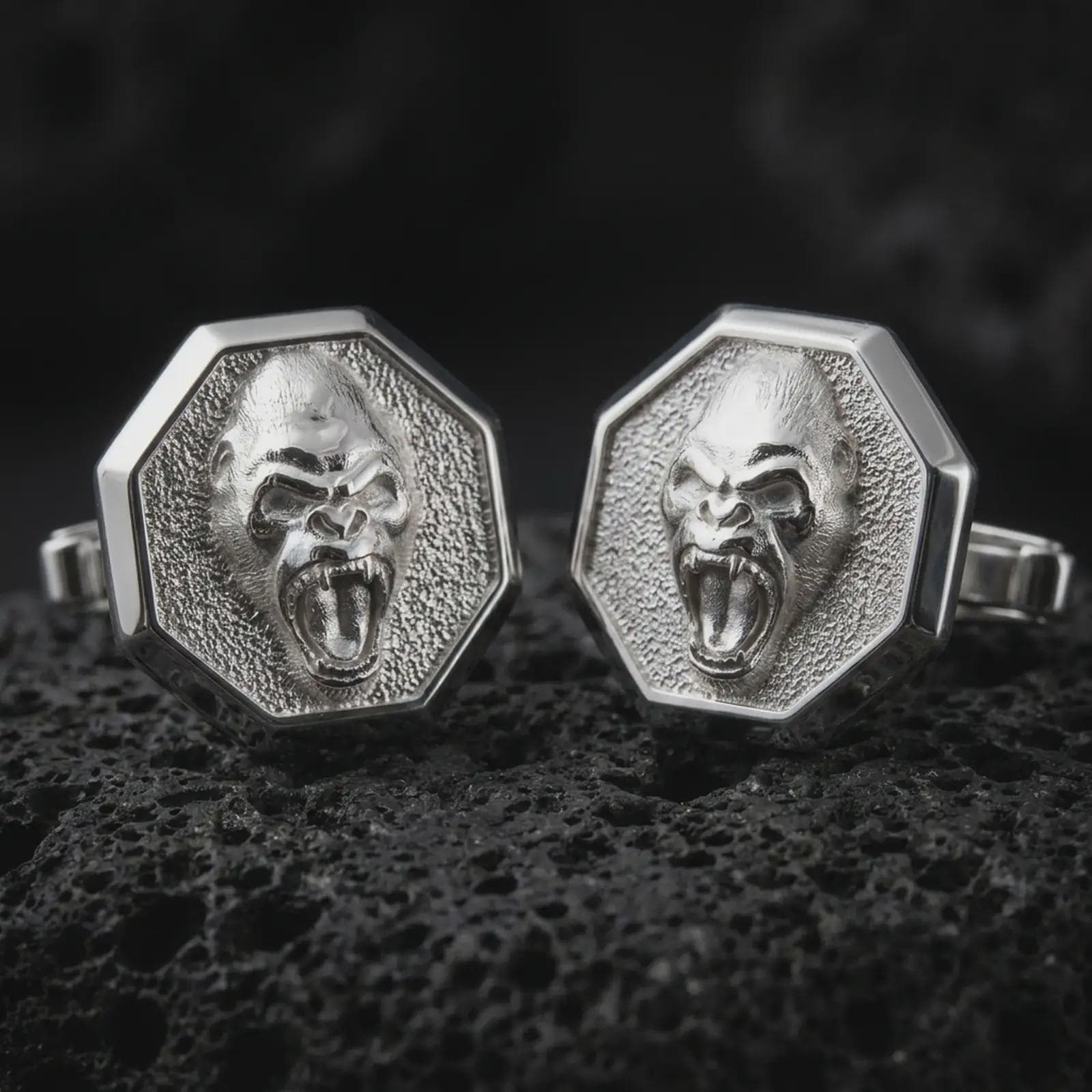 Polished silver gorilla cufflinks on textured volcanic rock, centered macro jewelry photo with detailed primate face sculpting and bright silver highlights.