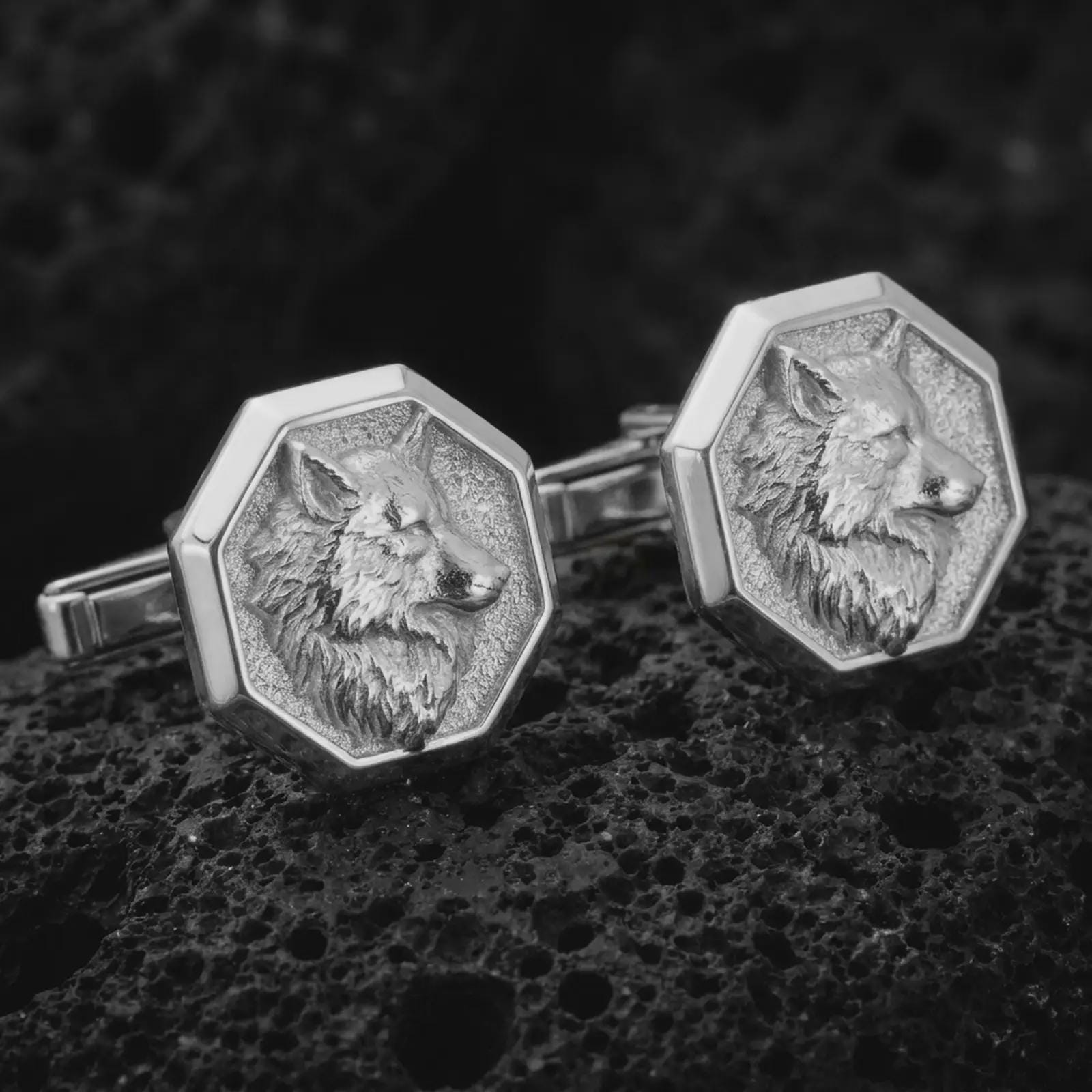 Polished silver wolf head cufflinks are displayed on black volcanic rock in a premium macro studio close-up with sharp facial texture and clean geometric edges.