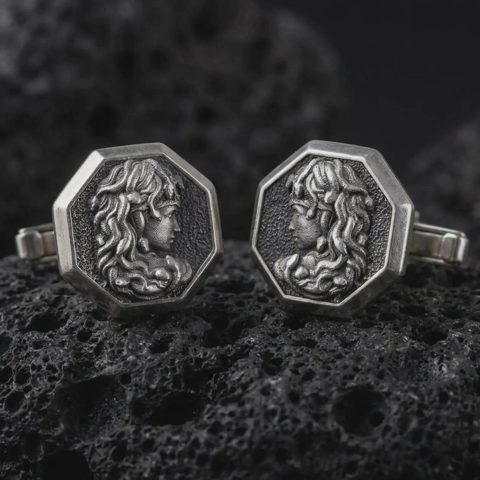 An oxidized Medusa cufflink pair rests on dark volcanic rock in a macro studio close-up, showing a sculpted Gorgon profile inside octagon frames.