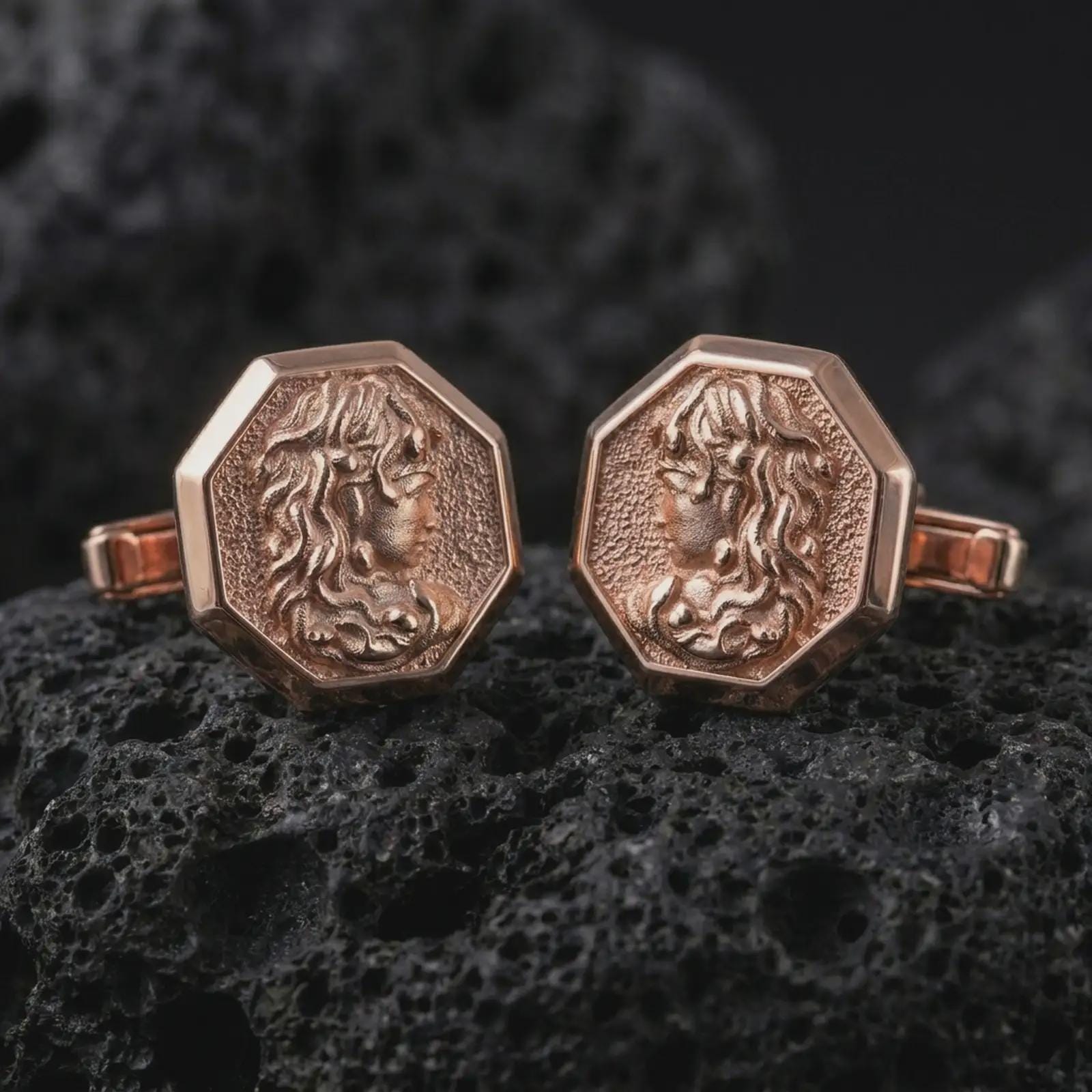 Rose gold plated Medusa cufflinks are photographed on porous volcanic rock in a close-up studio composition with refined luxury lighting.