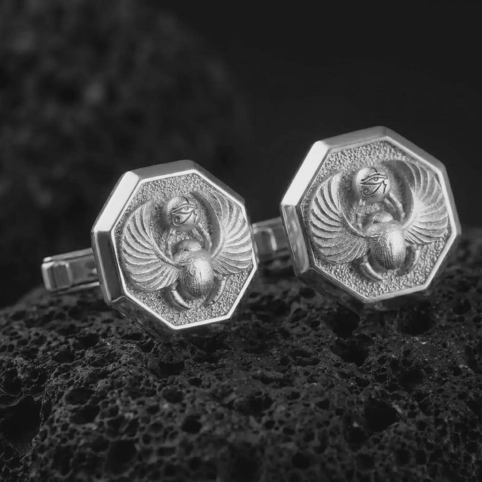 Polished silver scarab cufflinks are displayed on black volcanic rock in a premium macro studio close-up with detailed wings, beetle body, and Eye of Horus accent.