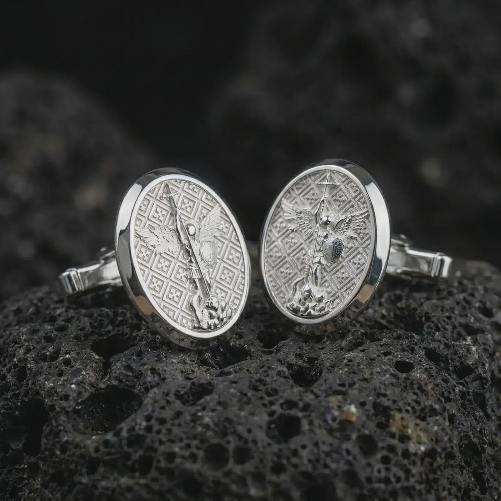 Polished silver Saint Michael cufflinks on textured volcanic rock, centered macro jewelry photo with detailed guardian angel medal engraving and bright silver highlights.