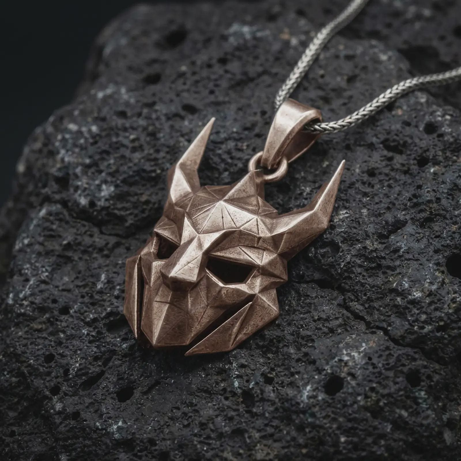 An Bronze Loki pendant rests on dark volcanic rock in a macro studio close-up, showing a faceted horned mask inspired by Norse mythology.