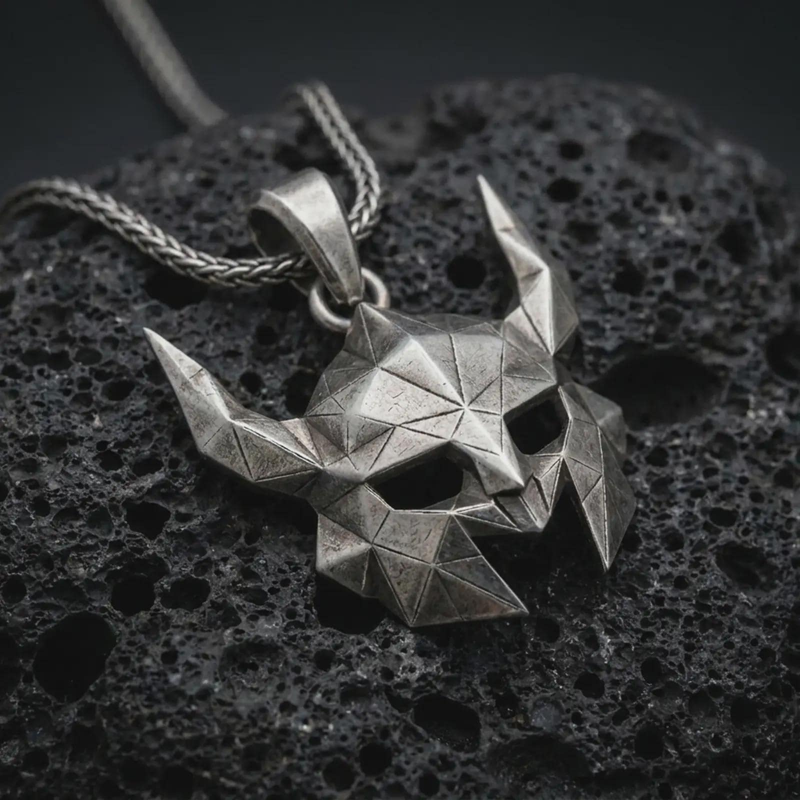 An oxidized Helm of Awe inspired mask pendant rests on dark volcanic rock in a macro studio close-up, showing a faceted horned Norse warrior design.