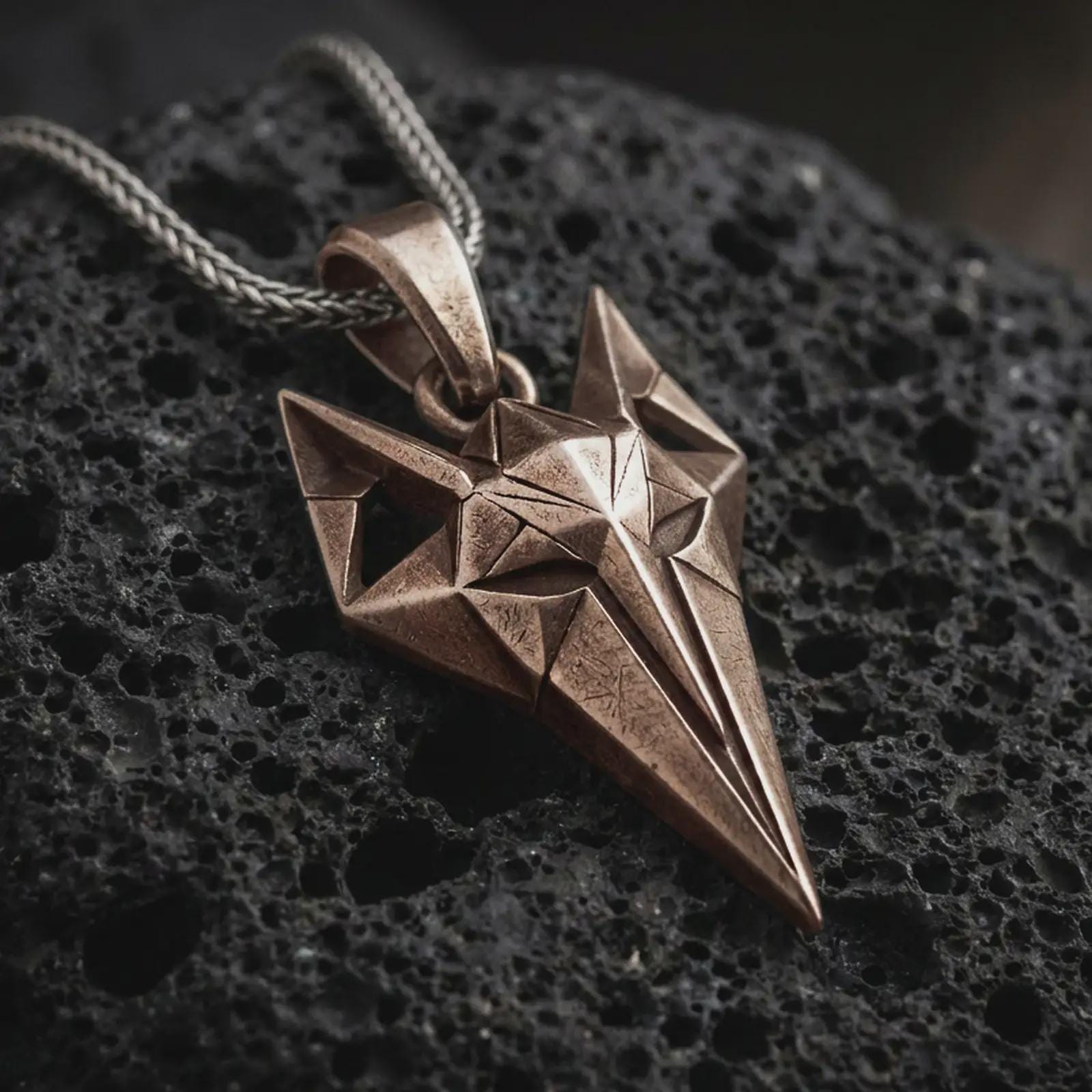 A bronze Freyja cat pendant rests on dark volcanic rock in a macro studio close-up, showing a faceted feline design inspired by Norse mythology.