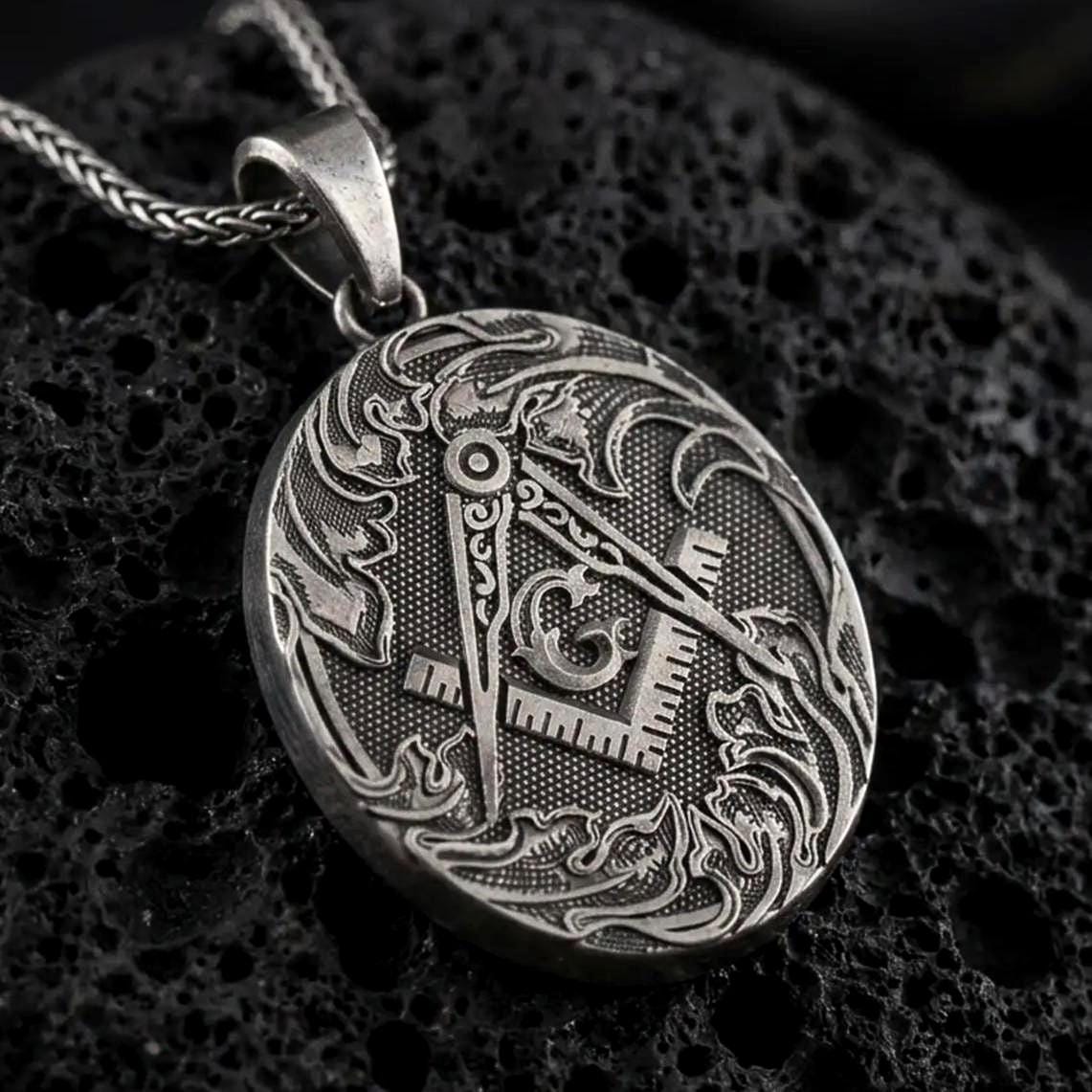 Oxidized Masonic square and compass pendant necklace on textured volcanic rock, close-up studio shot highlighting the Freemason symbol, ornate border, engraved details, and antique silver contrast.