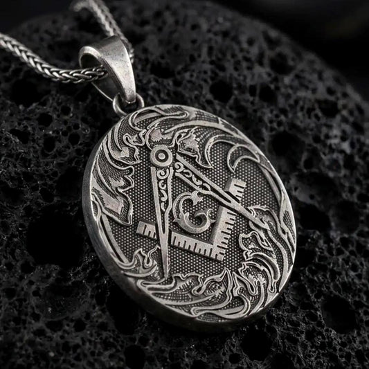 Oxidized Masonic square and compass pendant necklace on textured volcanic rock, close-up studio shot highlighting the Freemason symbol, ornate border, engraved details, and antique silver contrast.