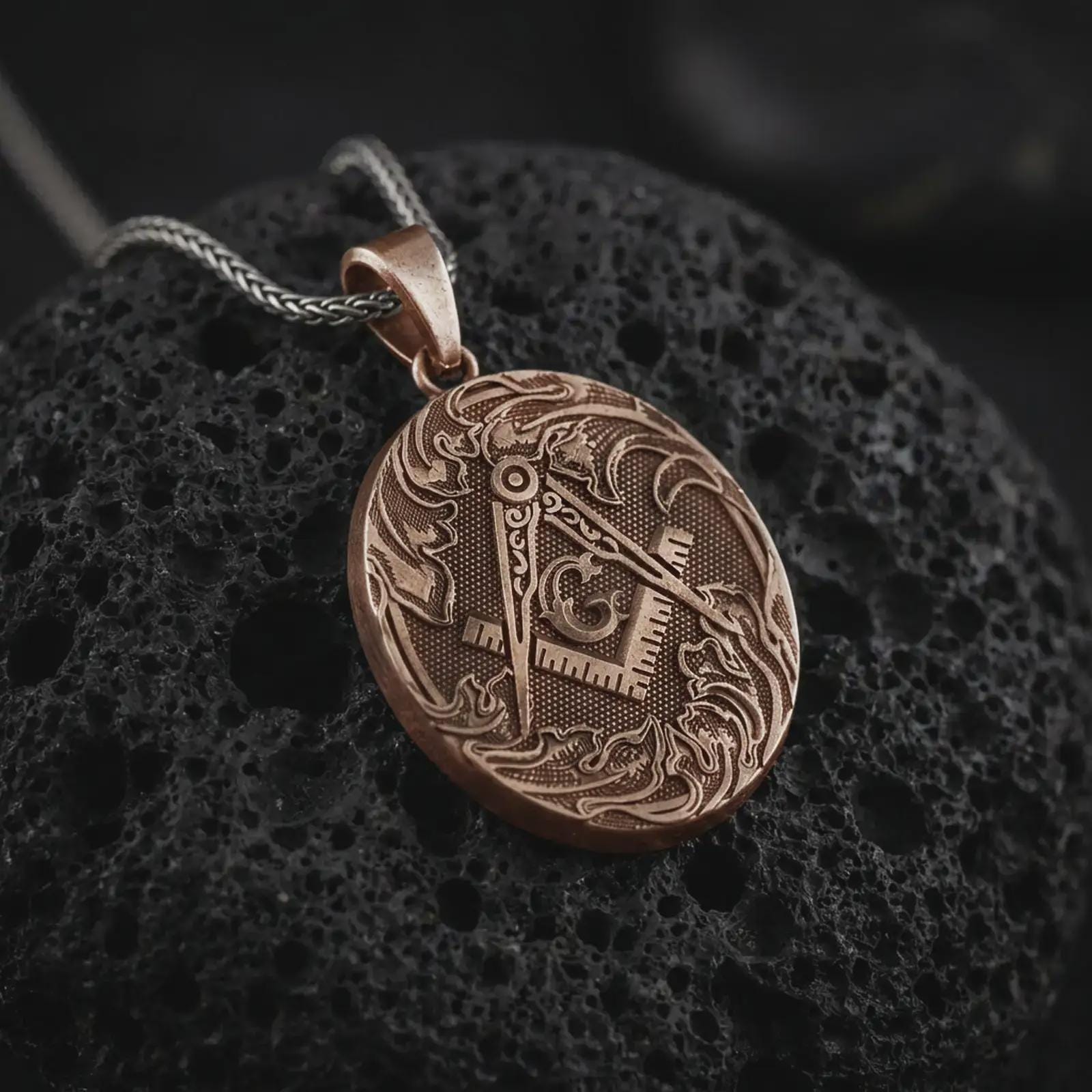 Bronze Masonic square and compass pendant necklace on textured volcanic rock, close-up studio shot highlighting the Freemason symbol, ornate border, engraved details, and antique silver contrast.