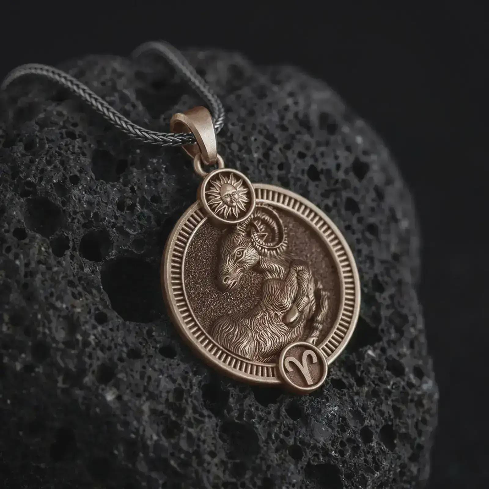 Bronze Aries zodiac pendant necklace on textured volcanic rock, close-up studio shot highlighting the ram relief, sun detail, Aries symbol, and antique silver contrast.