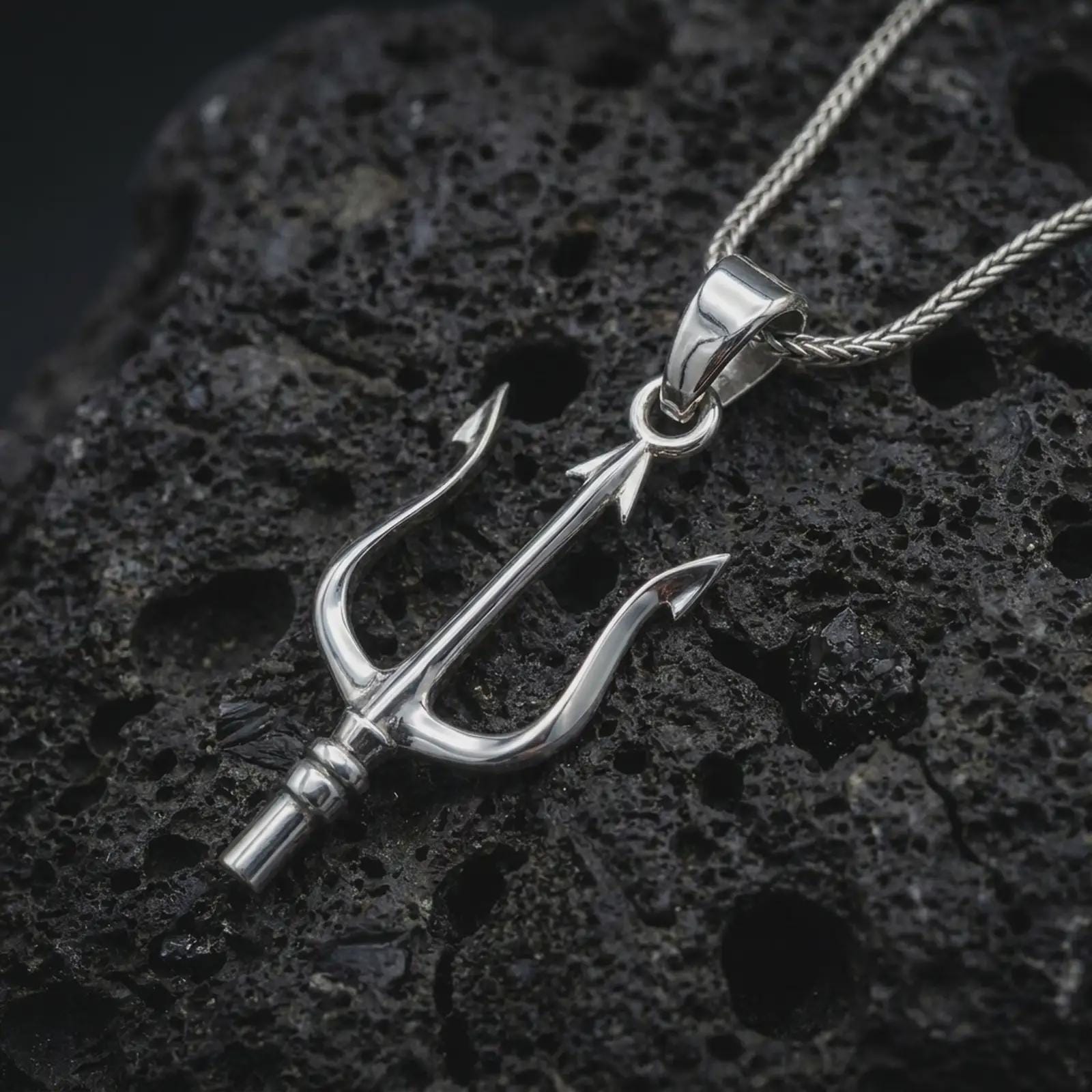Polished silver Poseidon trident pendant necklace on textured volcanic rock, centered macro jewelry photo with detailed sea god weapon design and bright silver highlights.