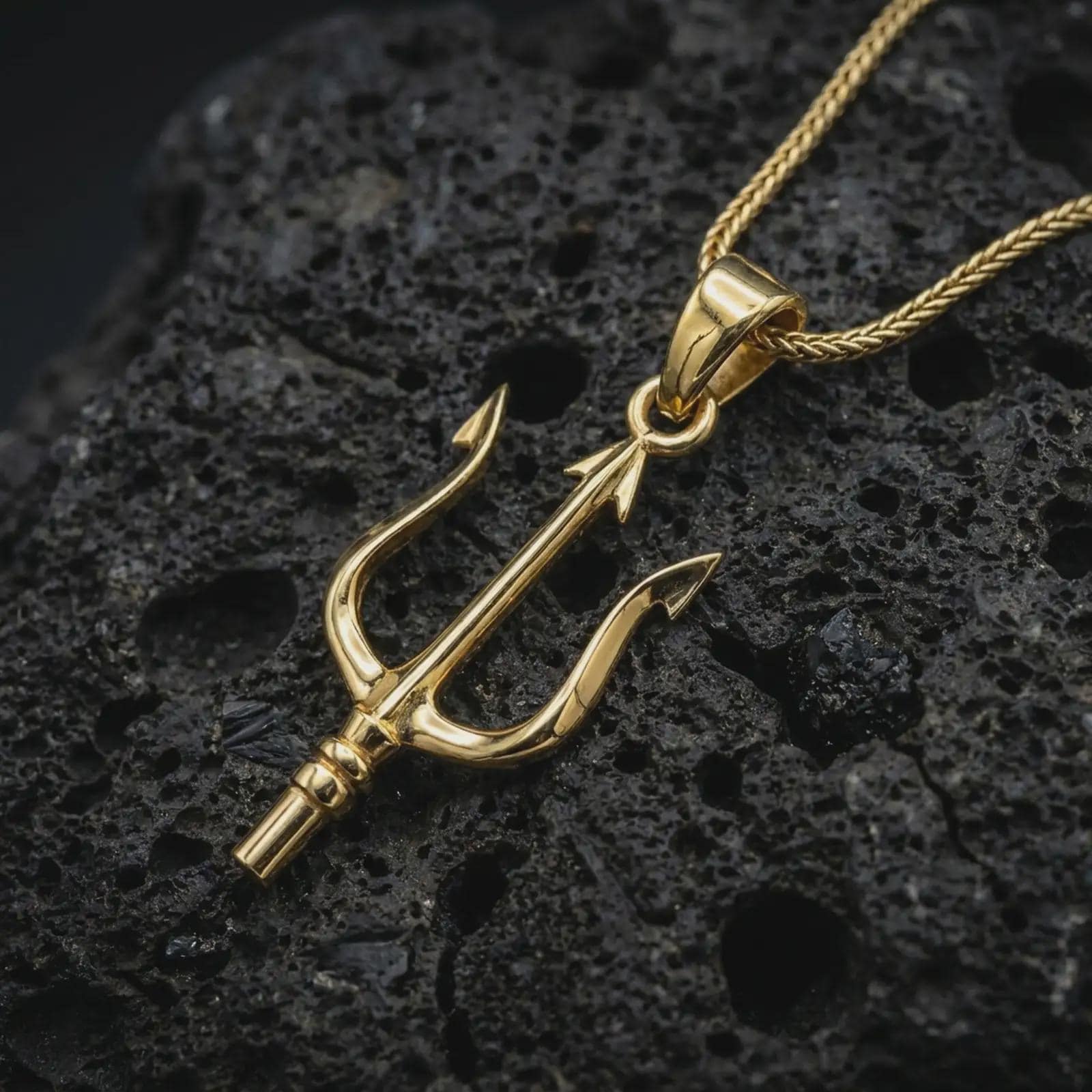 Gold plated Poseidon trident pendant necklace on textured volcanic rock, premium close-up studio composition emphasizing the Greek mythology trident, sculpted prongs, and bold pendant silhouette.